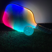 PG RGB pickleball paddle glowing blue, pink, cyan and amber on turf, with a perforated ball in front.